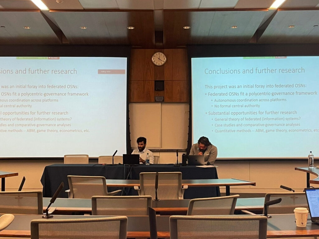 SpectrumX Postdoc Ali Palida presenting "Governance of Federated Platforms" with Will Rinehard, from the Center for Growth and Opportunity from Utah State University. Provided.
In the image, Ali and Will are at laptops at the front of the room in front of presentation screens.