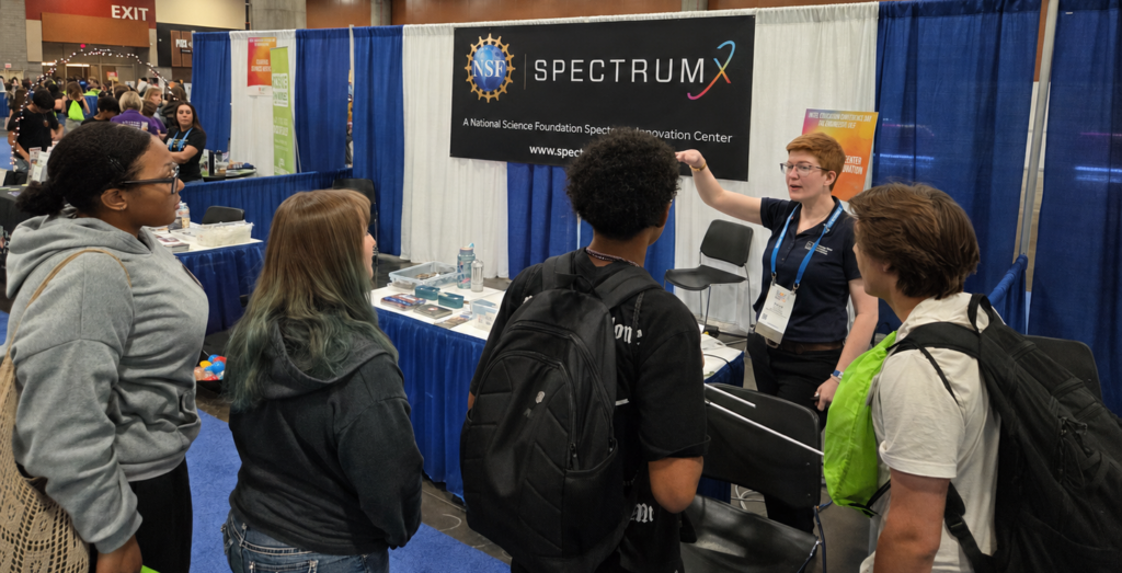 An NRAO staff member with short red hair and pale skin speaks to four students at a science convention