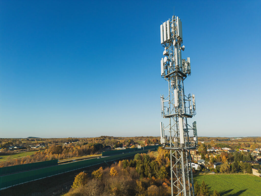 Cellular antenna in rural environment (lots of trees and open space visible). There is a clear, blue sky, and the trees' leaves are turning orange and brown.