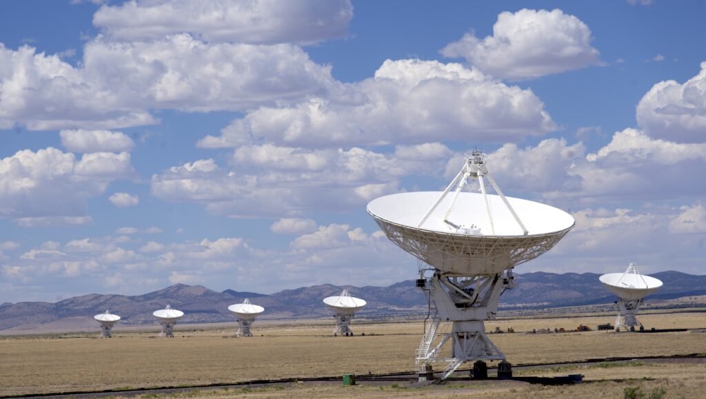 Photo of large white radio antennas. One is in the forefront. Five appear behind. Antennas are located in the dessert in front of a range of mountains