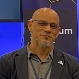 Headshot of Rafael Rodriguez Solis--a bald man with tan skin, a gotee, and glasses smiles. He wears a black suit jacket with a lapel pin and a gray button-up shirt with a lanyard.